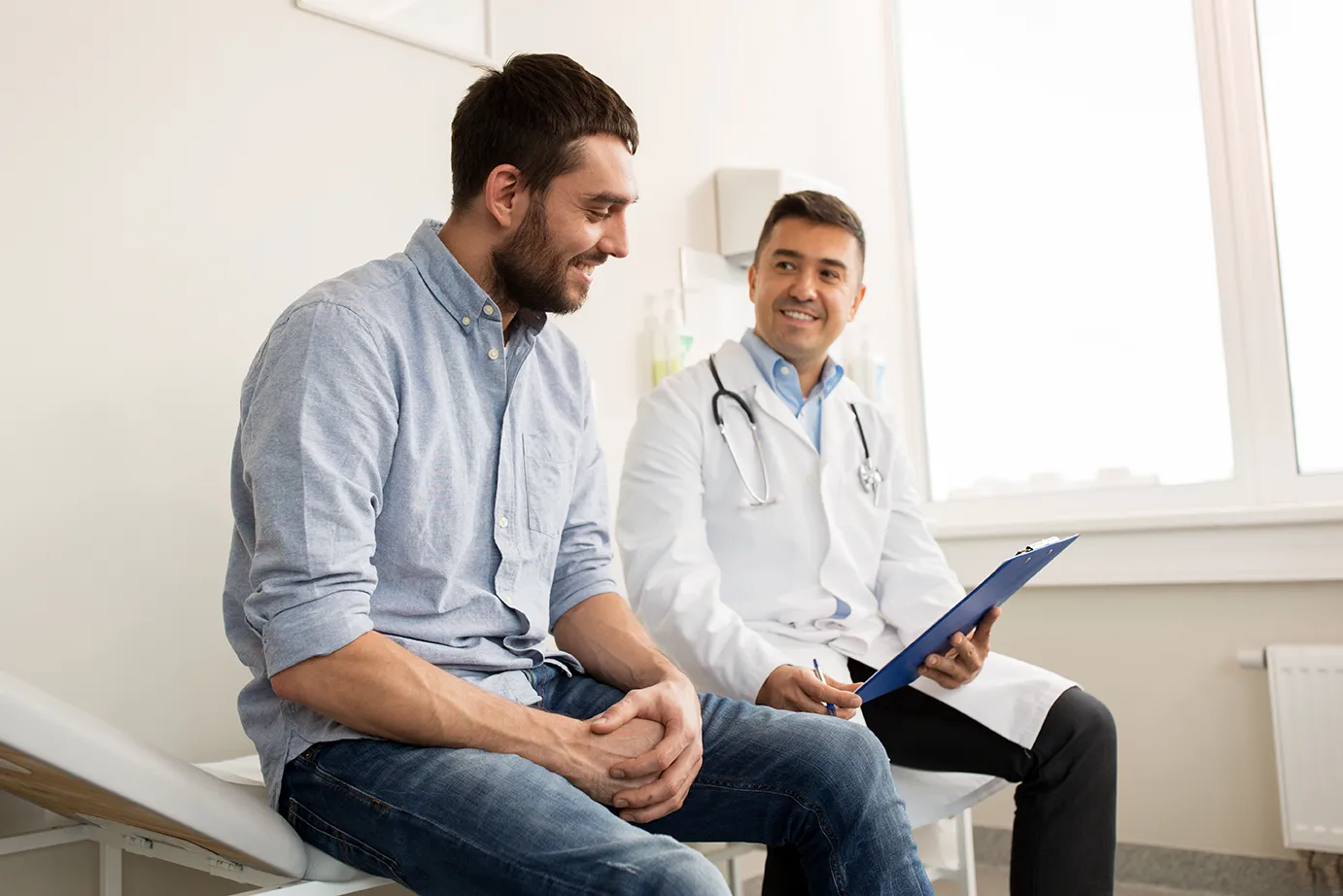 Smiling doctor in white coat with stethoscope talks to smiling patient on exam table, holding clipboard.
